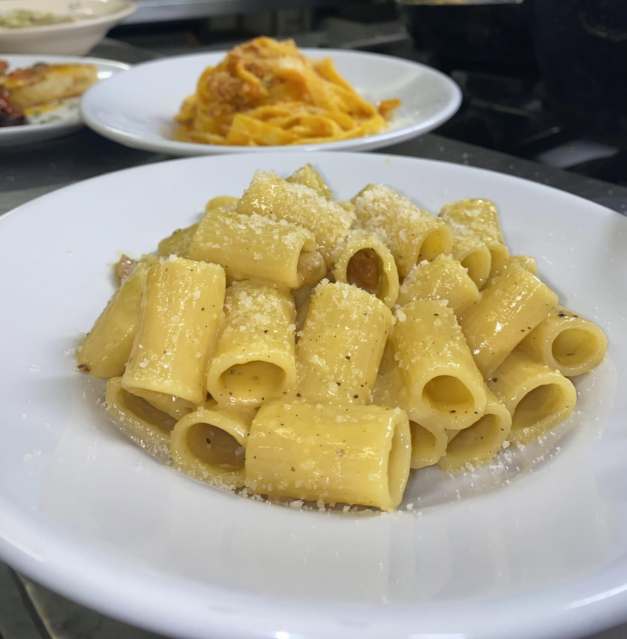 Rigatoni con formaggio grattugiato e pepe nero in piatto bianco, dietro tagliatelle al sugo di La Cicala e la Formica