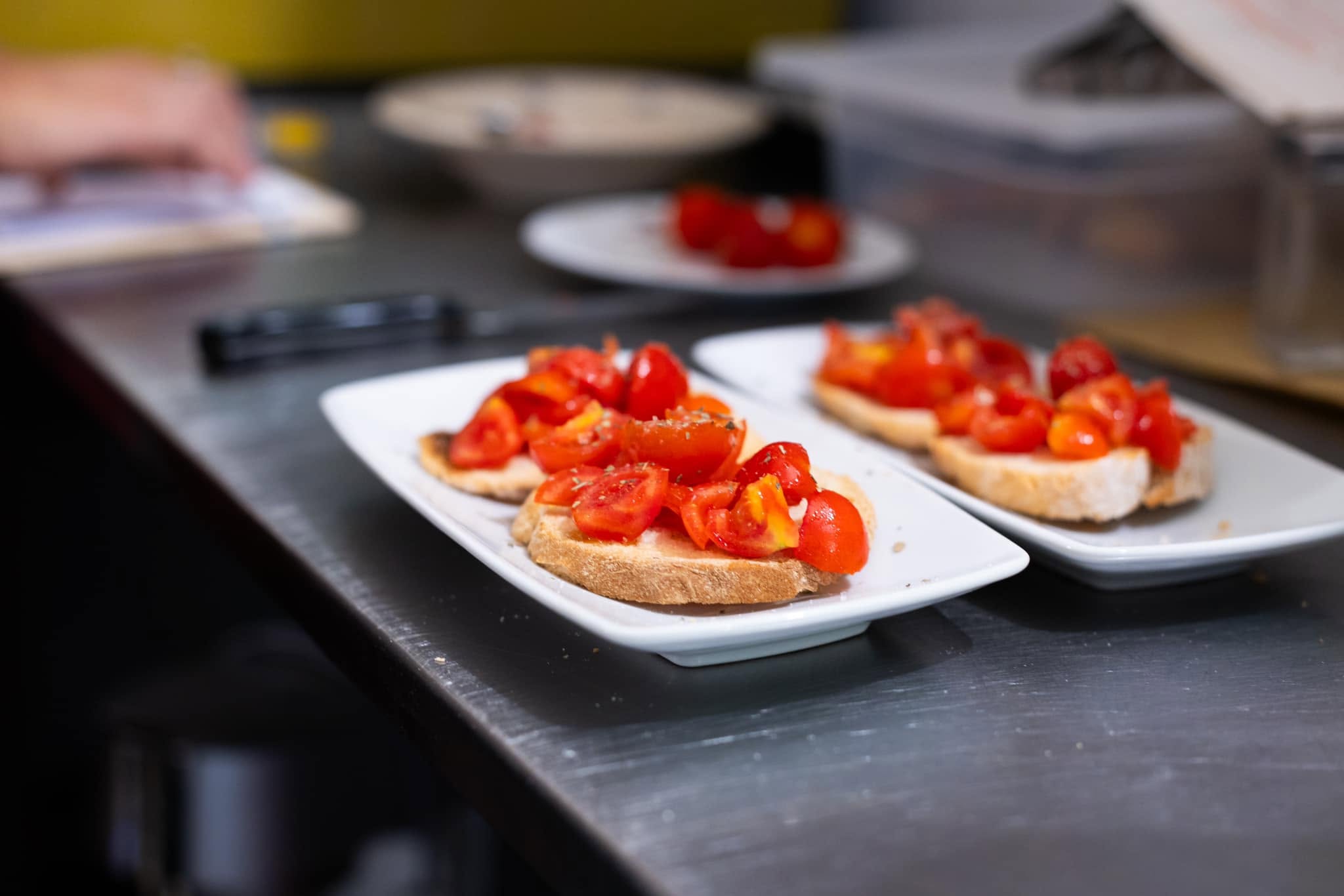 Bruschette con pomodorini tagliati e origano su piatti bianchi rettangolari, banco in acciaio della cucina La Cicala e la Formica