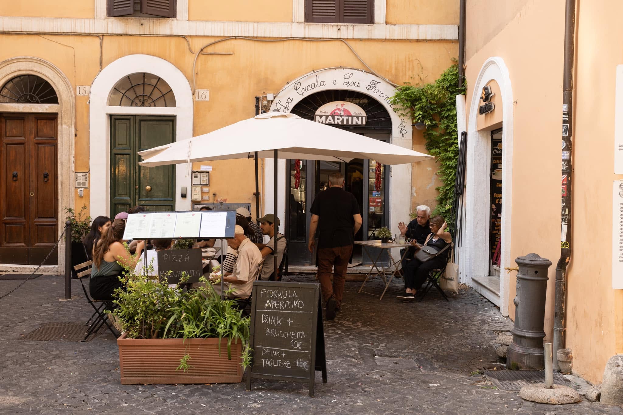 Dehors de La Cicala e la Formica: tavolini sotto ombrellone bianco su sampietrini, lavagna «Formula aperitivo», nasone