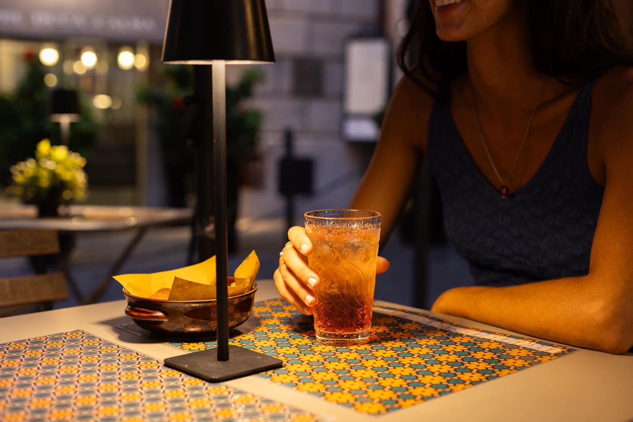 Donna al tavolo del dehors serale con bicchiere di spritz, ciotola di stuzzichini, lampada nera e tovaglietta a maioliche
